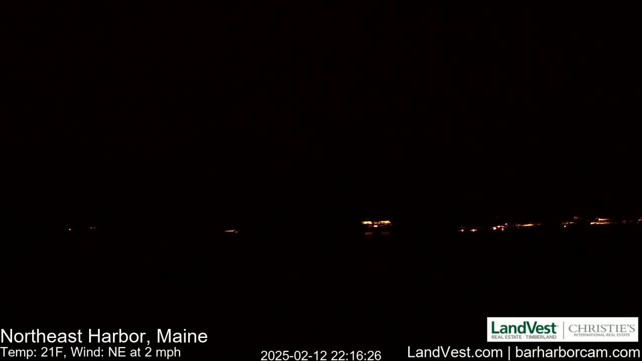 A nighttime, low-light image shows a distant line of faint orange lights, likely from a town or village, under a dark, clear sky with a northeast wind of 2 mph and a temperature of 21°F.