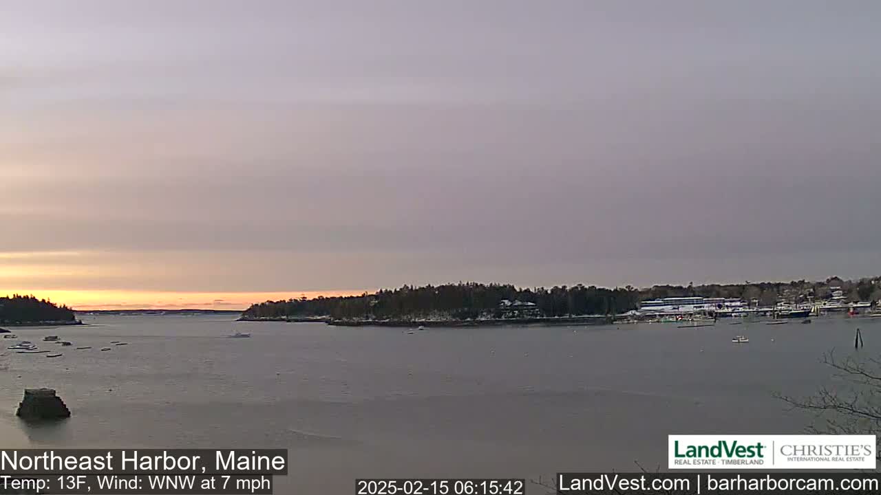 A calm harbor at sunrise with a partly cloudy sky and a temperature of 13°F, featuring a shoreline with snow-dusted trees and a few boats in the water.