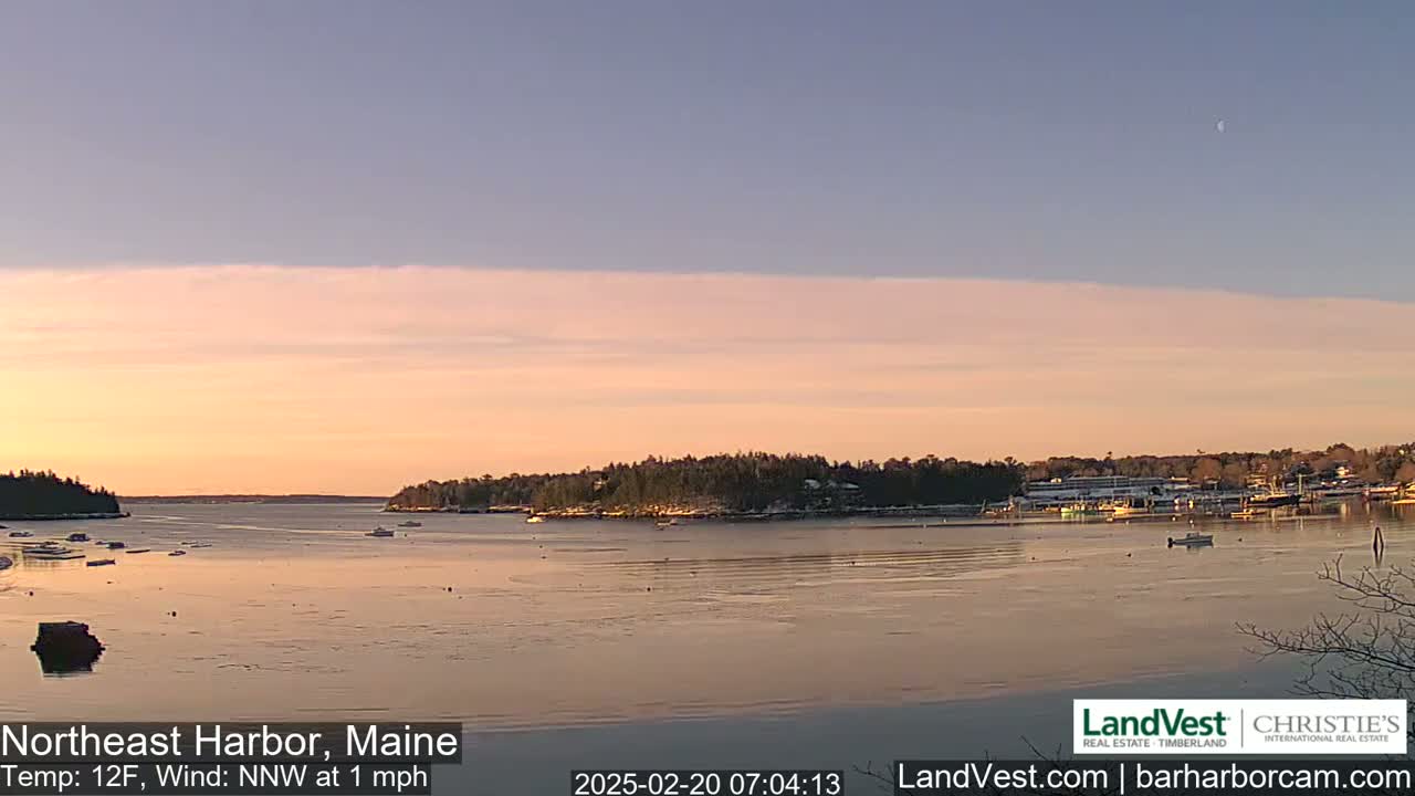 A calm harbor at sunrise with a few boats and a thin layer of ice on the water under a pale pink and blue sky.