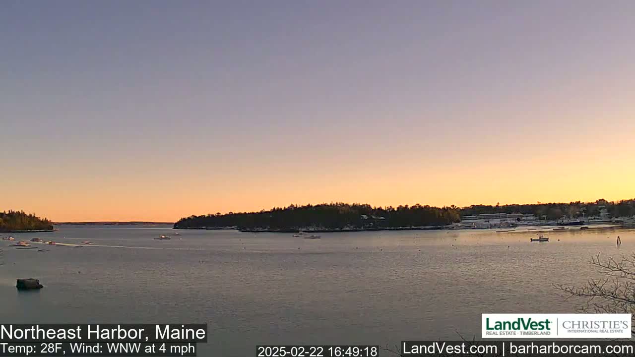 A calm body of water at sunset shows several boats and snow-covered land in the distance under a clear, pale orange and purple sky.