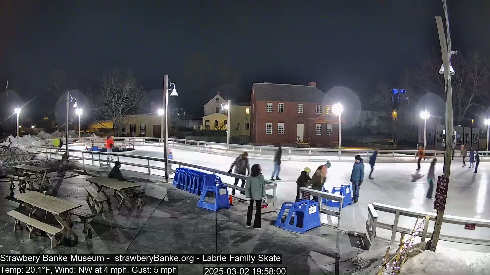 At night, under a clear sky with a northwest wind of 4 mph and gusts up to 5 mph, and a temperature of 20.1°F, people ice skate on an outdoor rink illuminated by several lights, near some picnic tables and buildings.