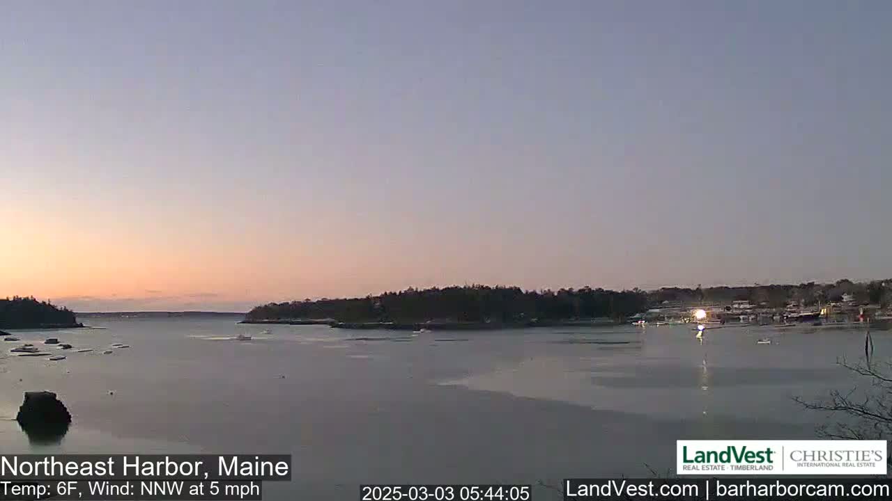 A calm harbor at sunrise with a partly icy surface, under a pale pink and purple sky, and a light north-northwest wind.