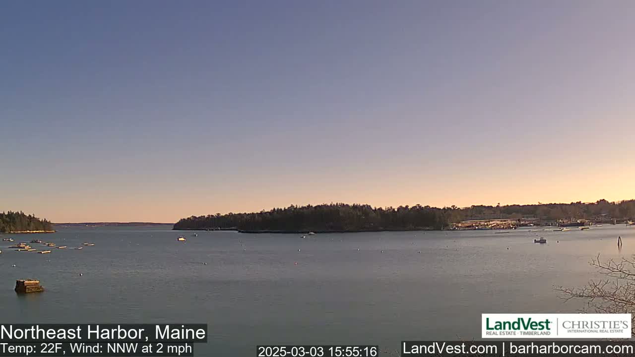 A calm harbor at sunset, with a few boats and wooded shorelines under a clear, pale sky.