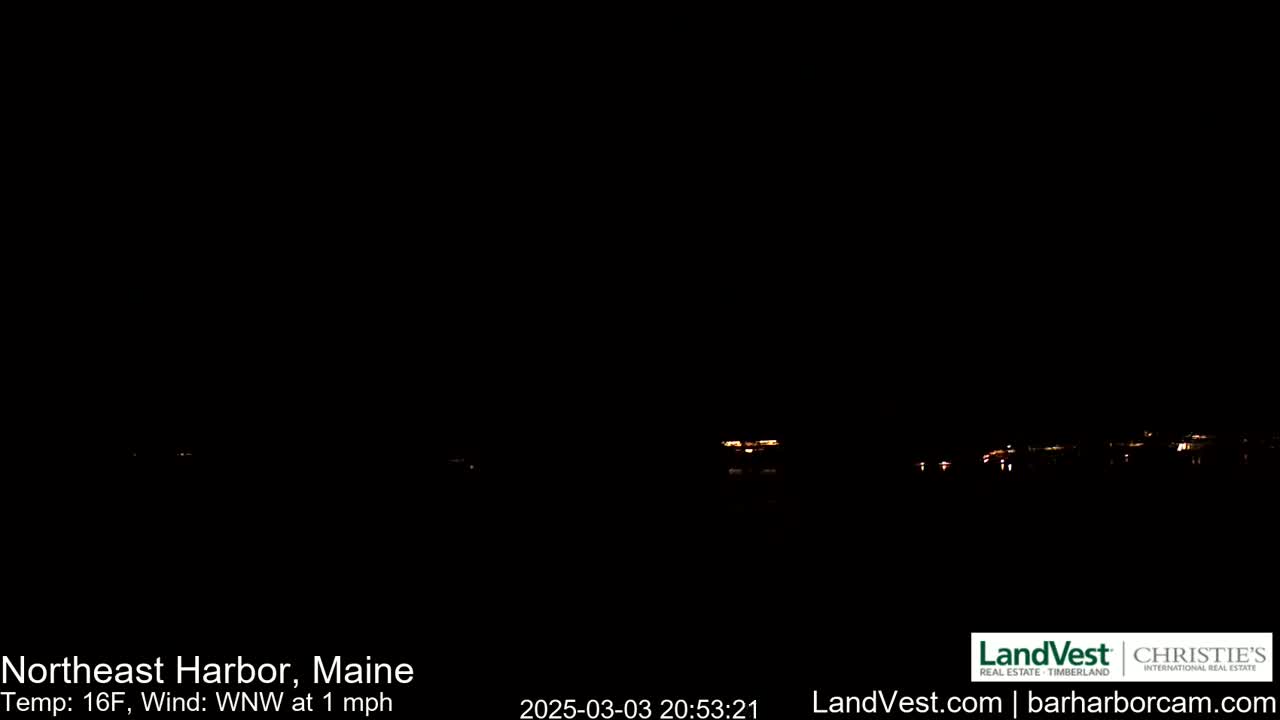 A nighttime long shot of dimly lit buildings across a dark body of water under a clear, dark sky with a 16°F temperature and a 1 mph wind from the west-northwest.