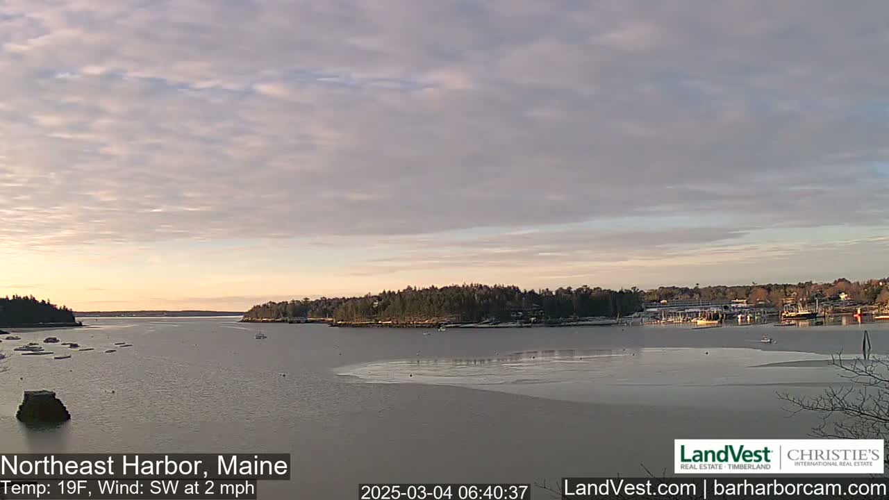 A partly cloudy sunrise over a calm harbor with some ice, showing a small town on the far shore.