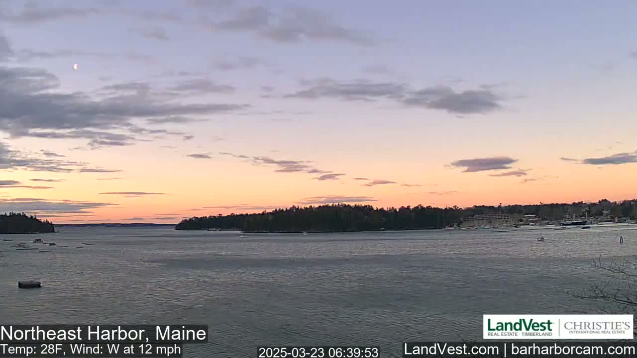 A calm body of water at sunset, under a partly cloudy sky with a crescent moon visible, shows a coastline with trees and a few buildings in the distance.