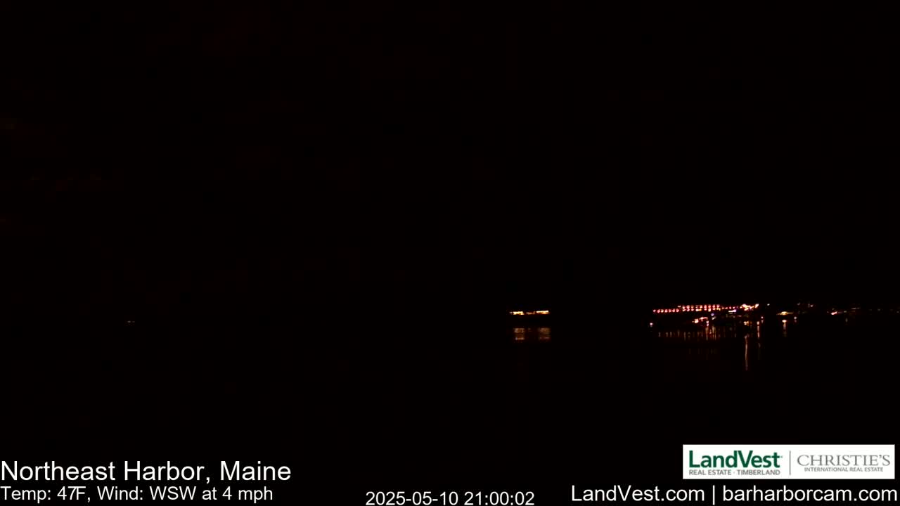 A nighttime view of dimly lit buildings across a dark body of water under a cloudy sky, with a light wind.