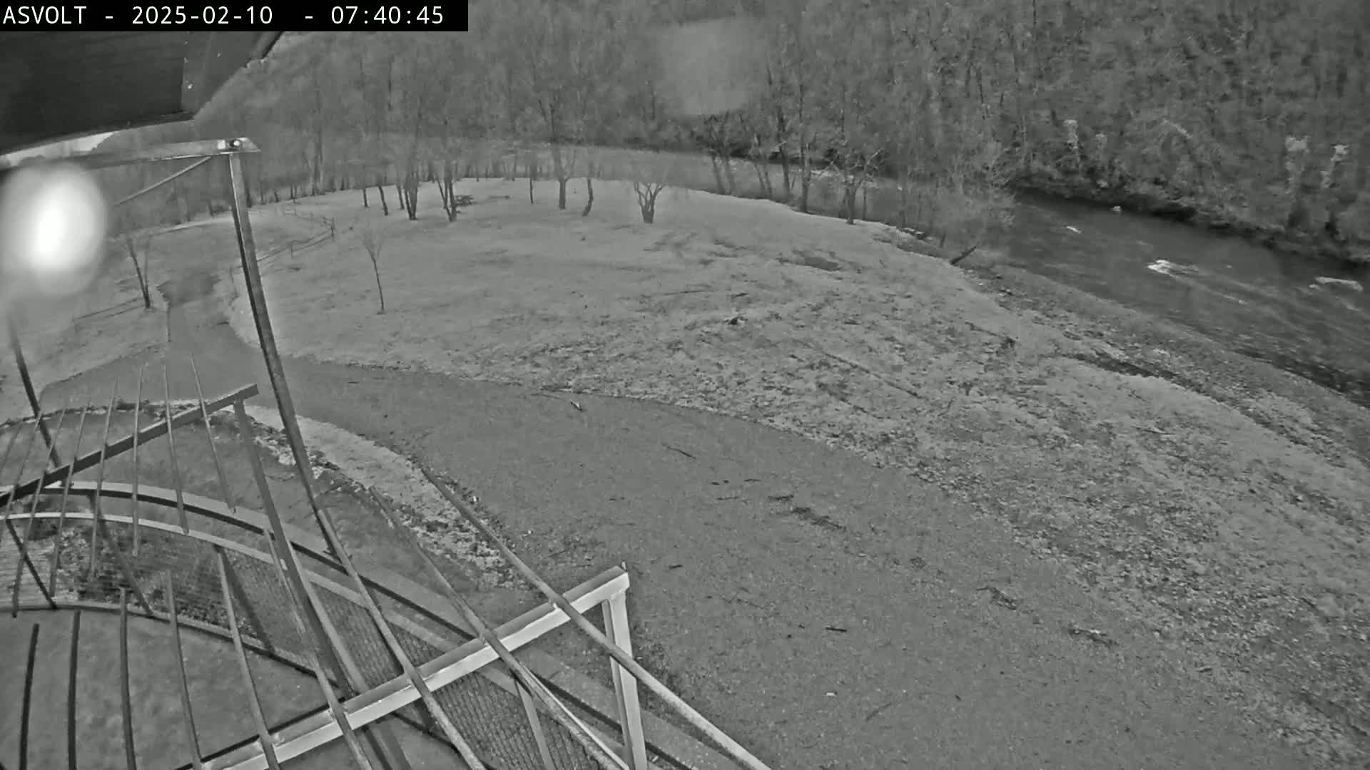 A high-angle grayscale nighttime view shows a gravel area next to a flowing river bordered by bare trees.