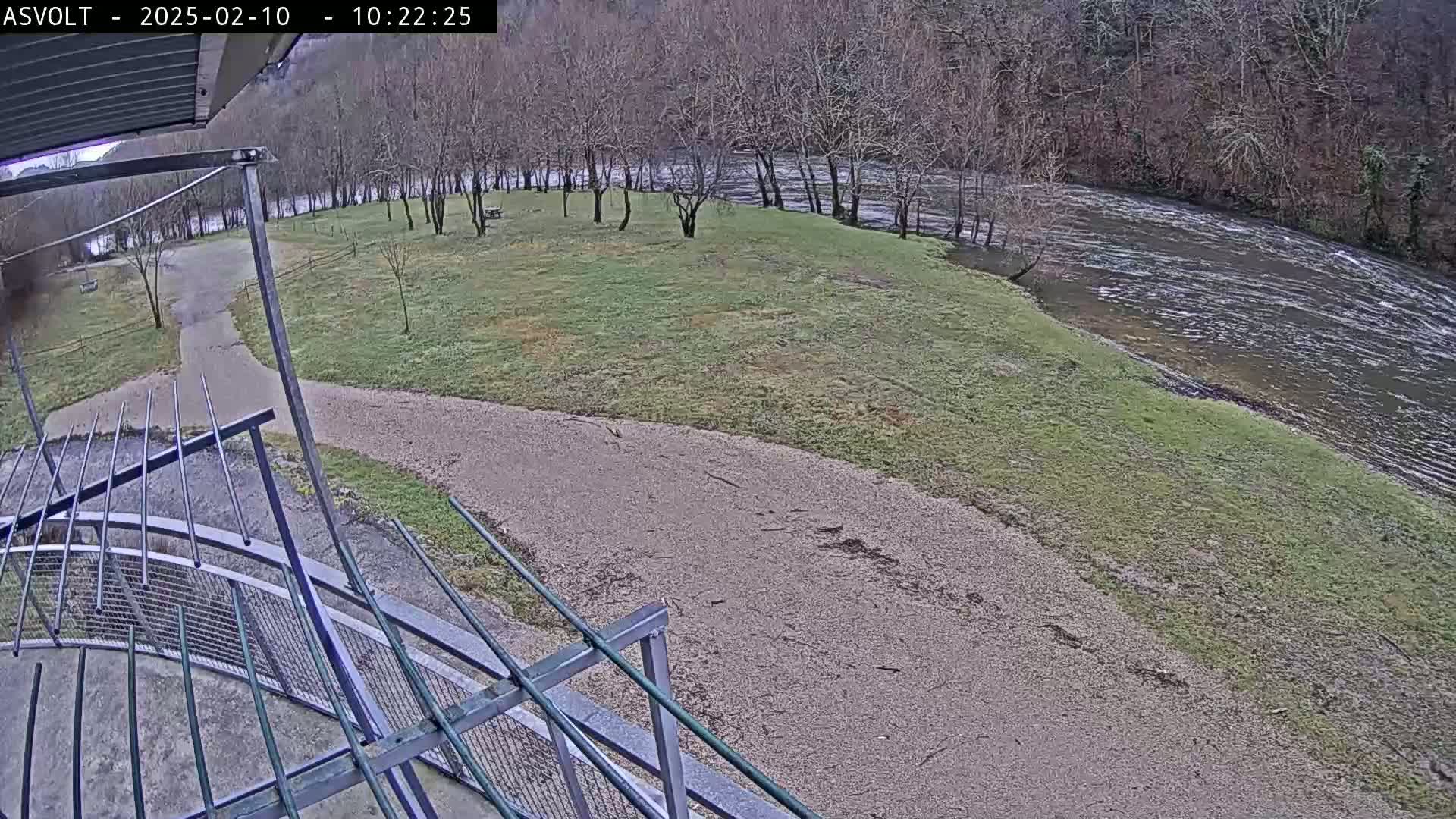 A gravel path leads to a grassy area beside a flowing river, surrounded by bare winter trees.