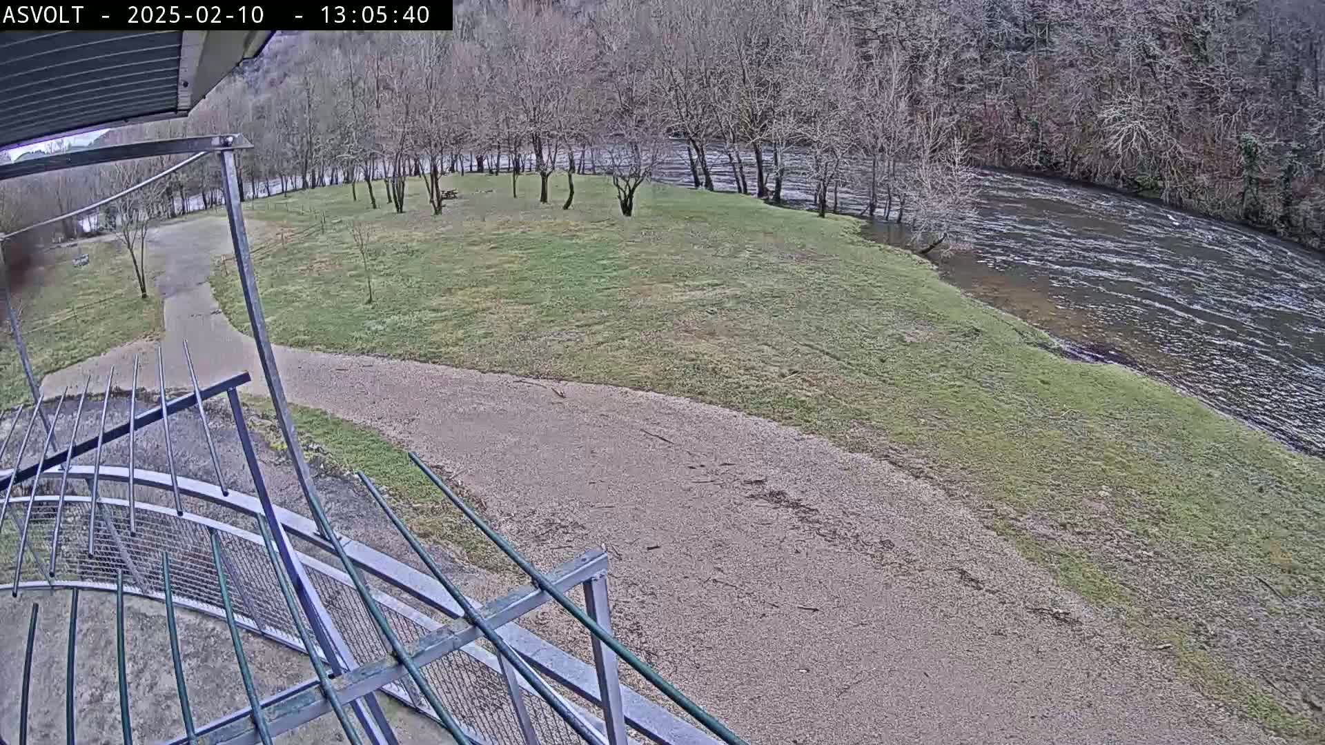 A gravel path leads to a grassy area beside a river under a cloudy sky, viewed from an elevated position behind a metal railing.