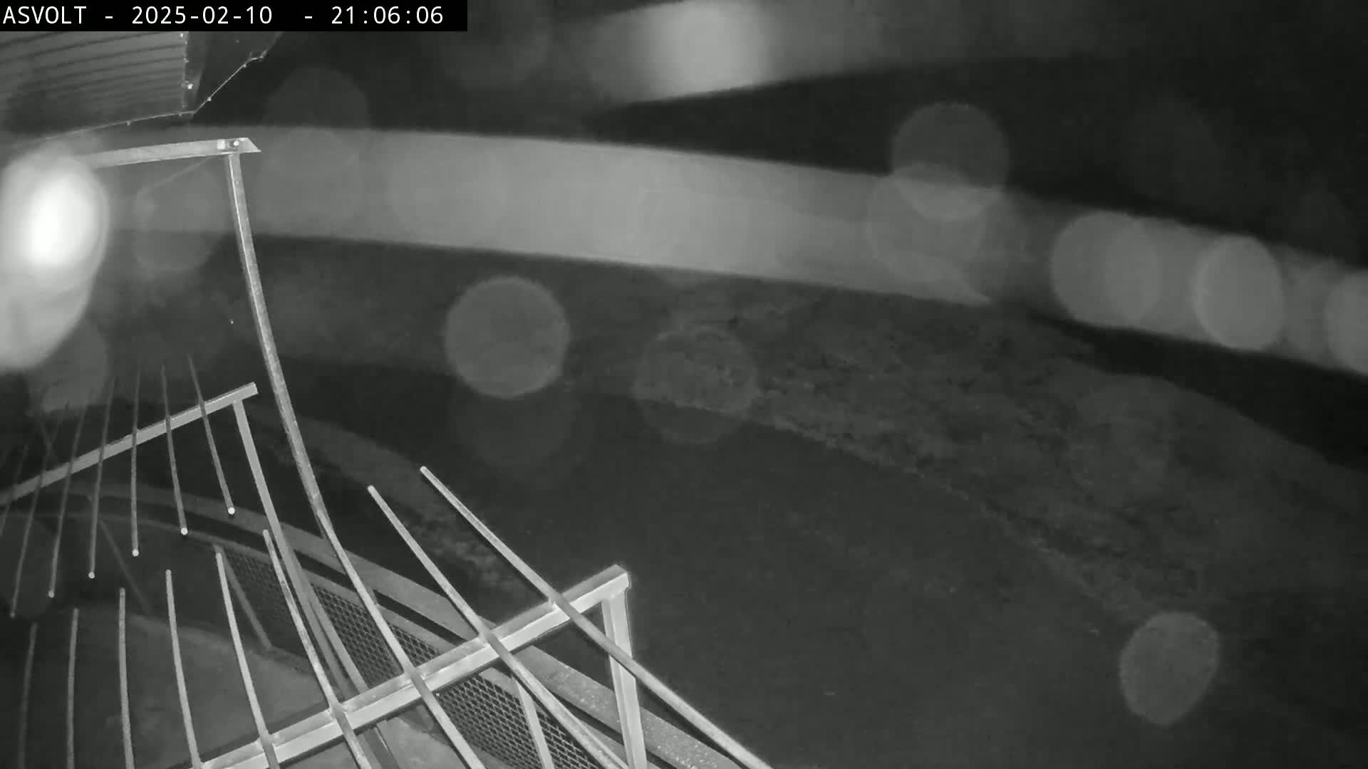 A nighttime, rainy view of a metal railing overlooking a dark, ground-level area.