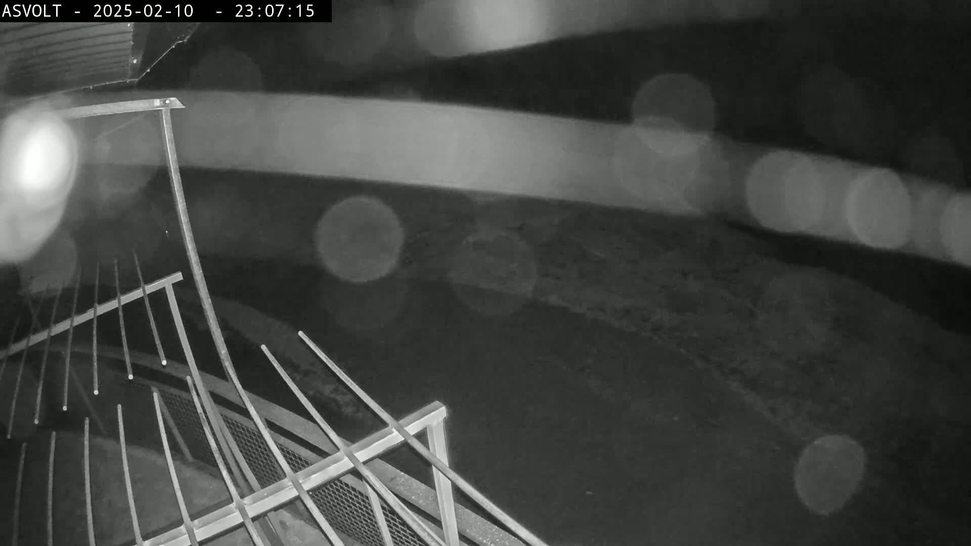 A nighttime, rainy view from an elevated position shows a metal railing and a dark, blurry ground below.