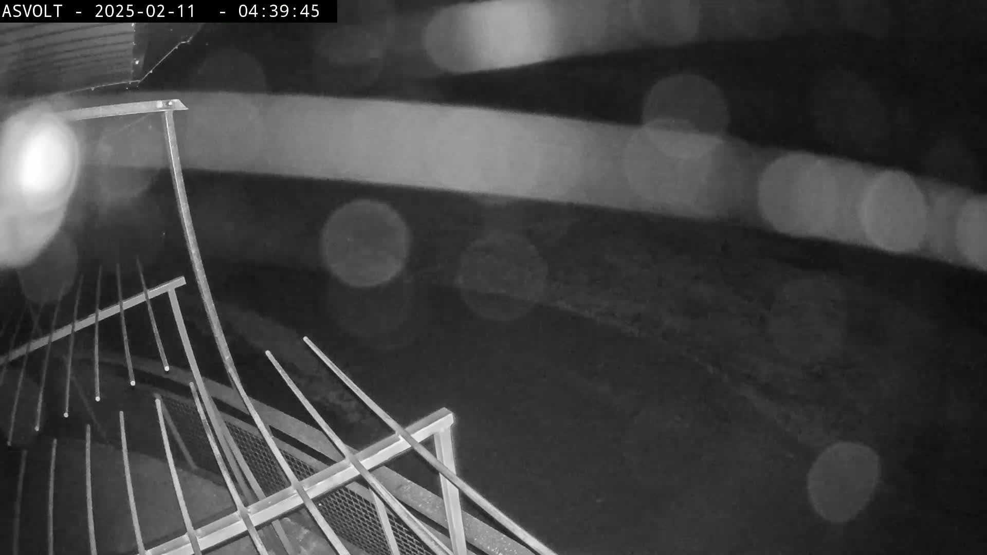 A nighttime, rainy view from an elevated position shows a metal railing and blurred background.
