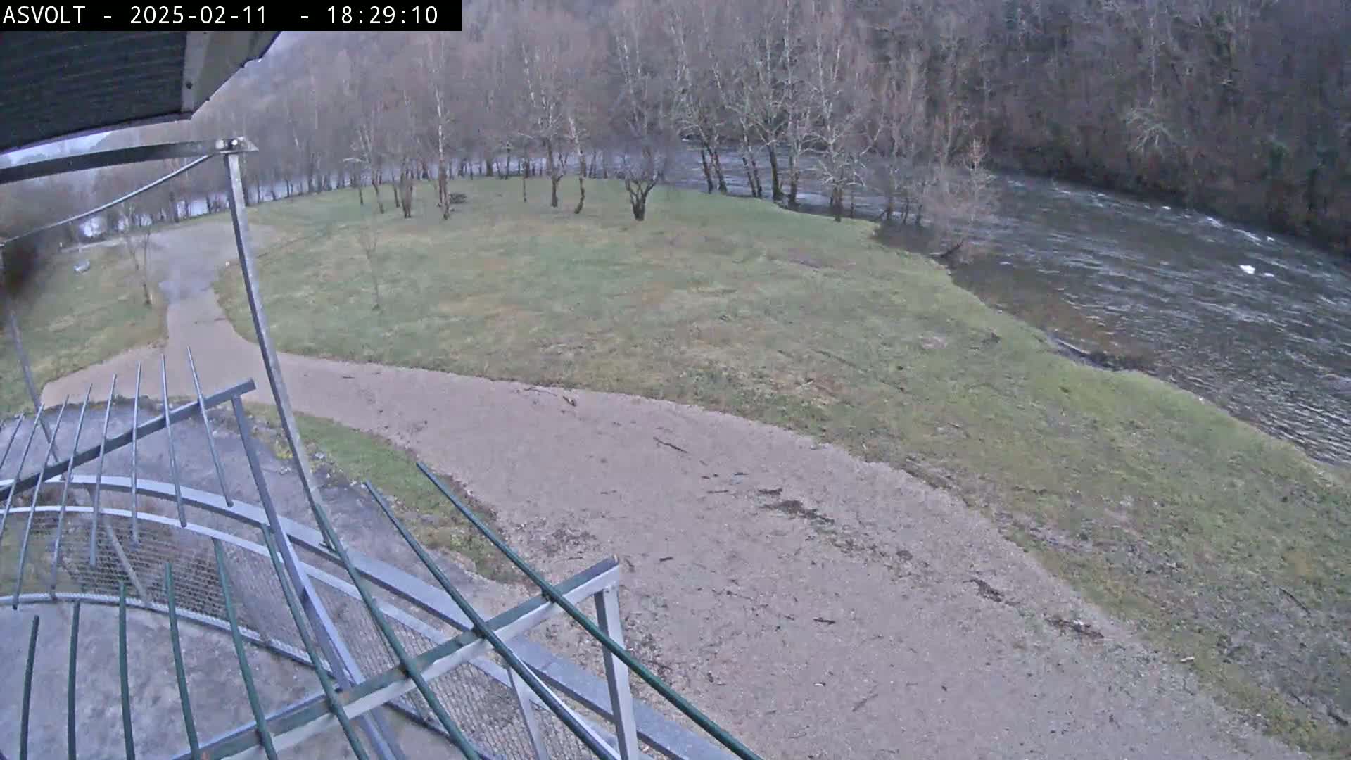 A gravel path leads to a grassy area beside a flowing river, with bare trees lining the riverbank in overcast conditions.