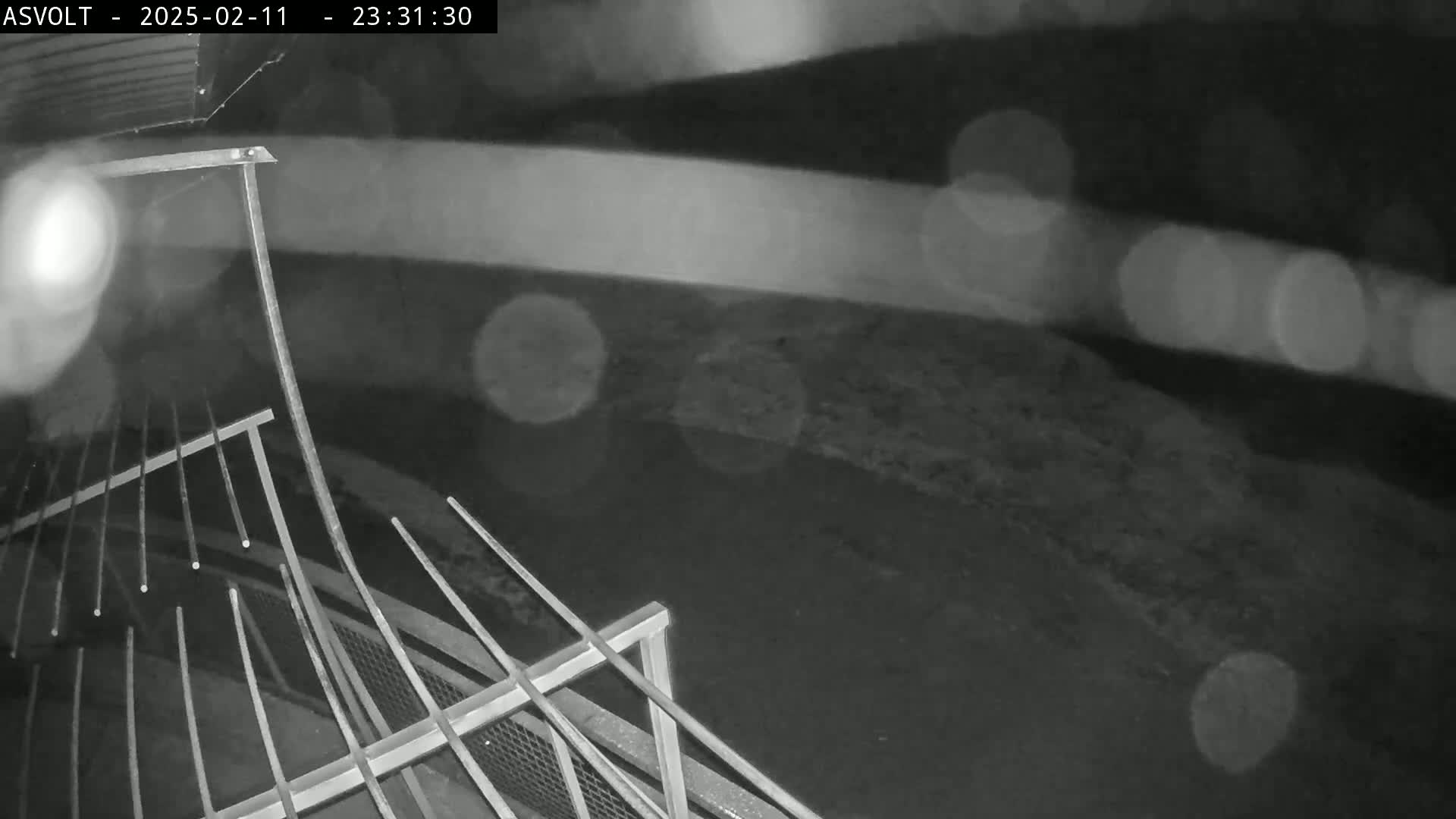 A nighttime, rainy view shows a section of metal railing, with blurred lights in the background.