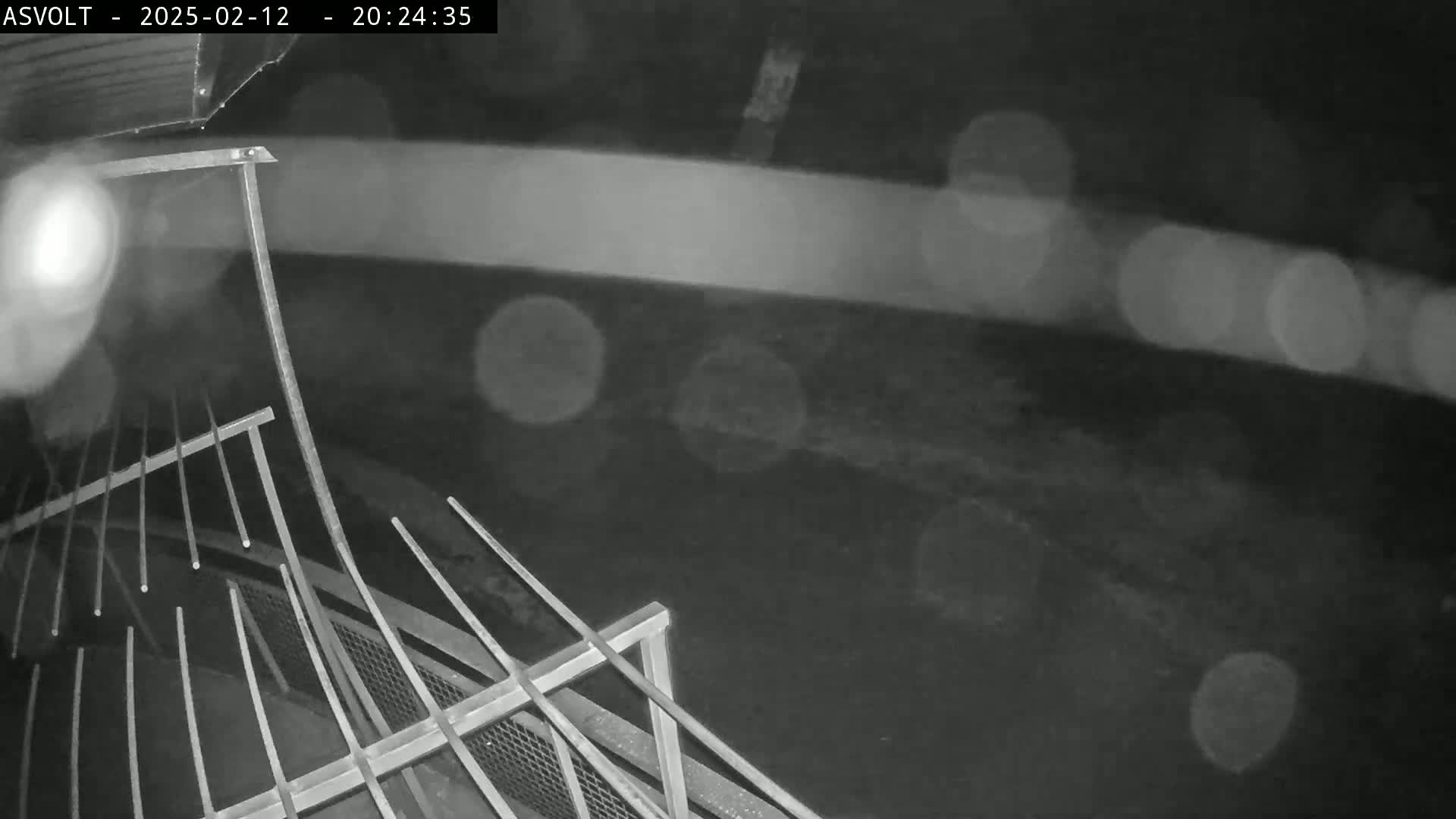 A nighttime, rainy view of a metal railing overlooking a dark, blurry area.