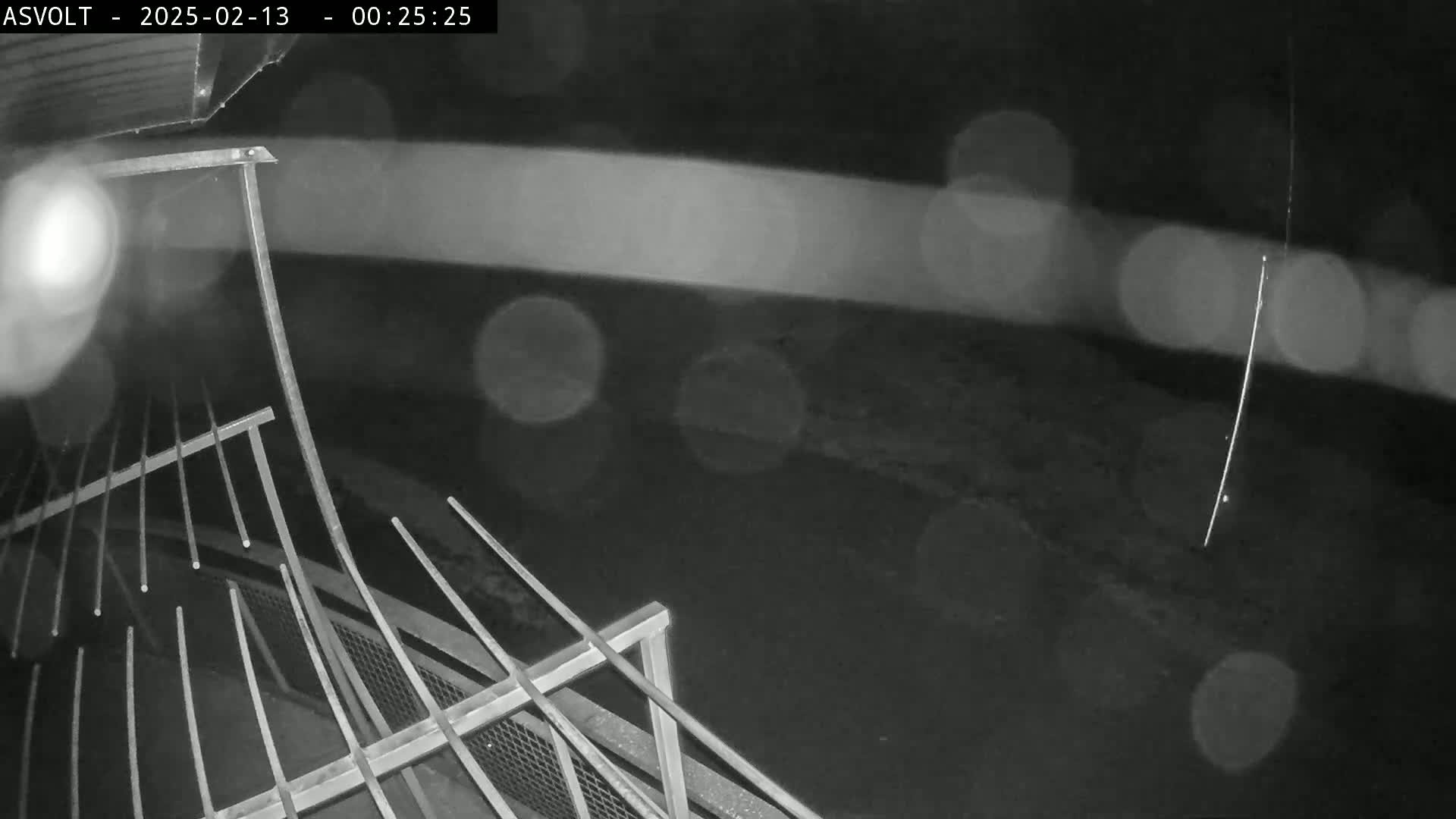 A nighttime, rainy view shows a section of metal railing overlooking a dark, blurry area.