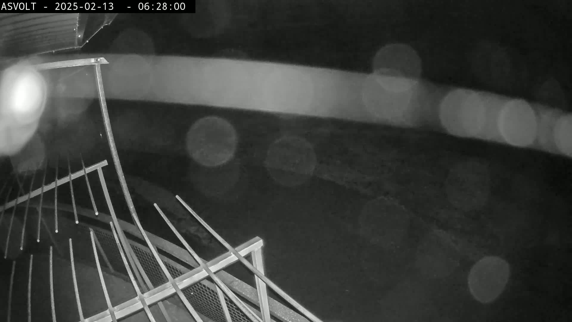 A nighttime, rainy view shows a metal railing in the foreground with blurred lights in the background.