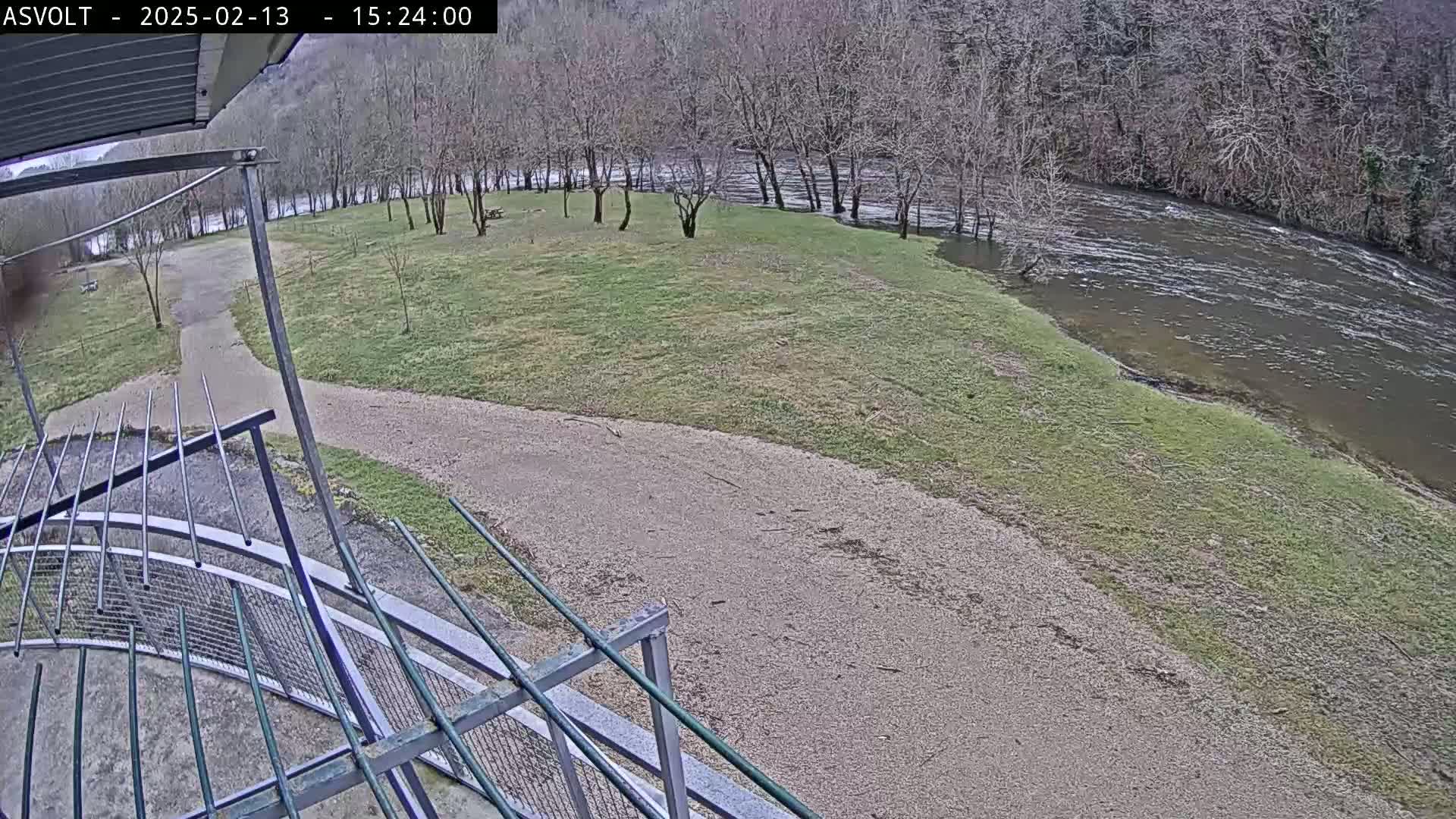 A gravel path leads to a grassy area beside a river, where bare trees line the bank under an overcast sky.