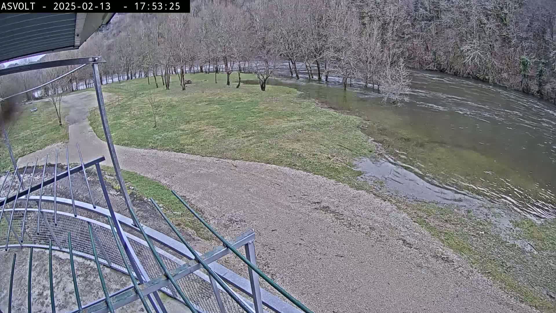 A gravel path leads to a grassy area beside a river that has overflowed its banks, partially flooding the land under an overcast sky.