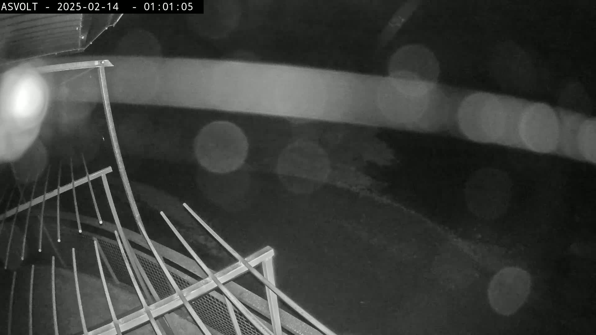 A nighttime, rainy view shows a section of metal railing and a dark ground below.