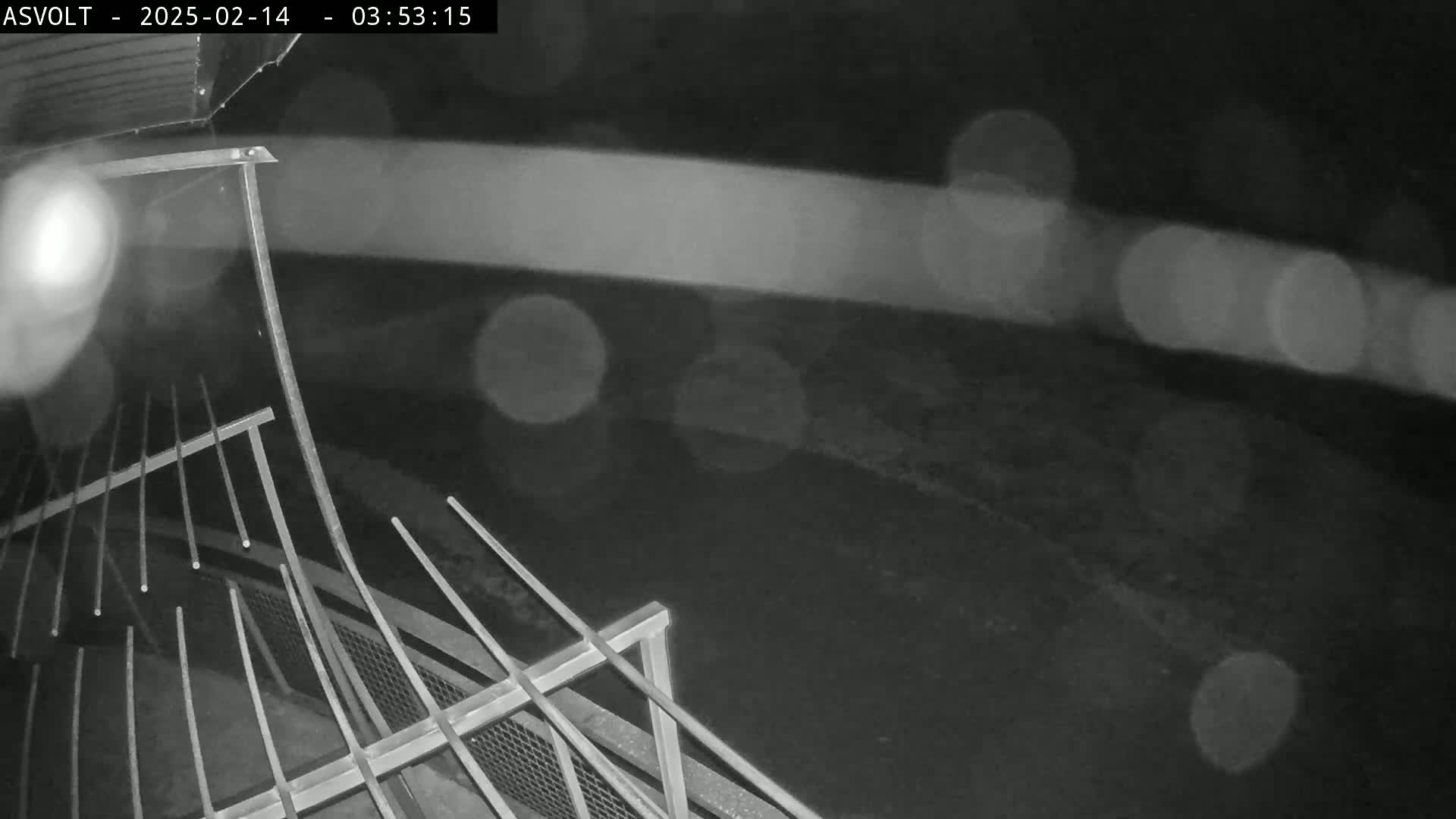 A nighttime, rainy view from an elevated vantage point shows a metal railing and blurred lights in the distance.