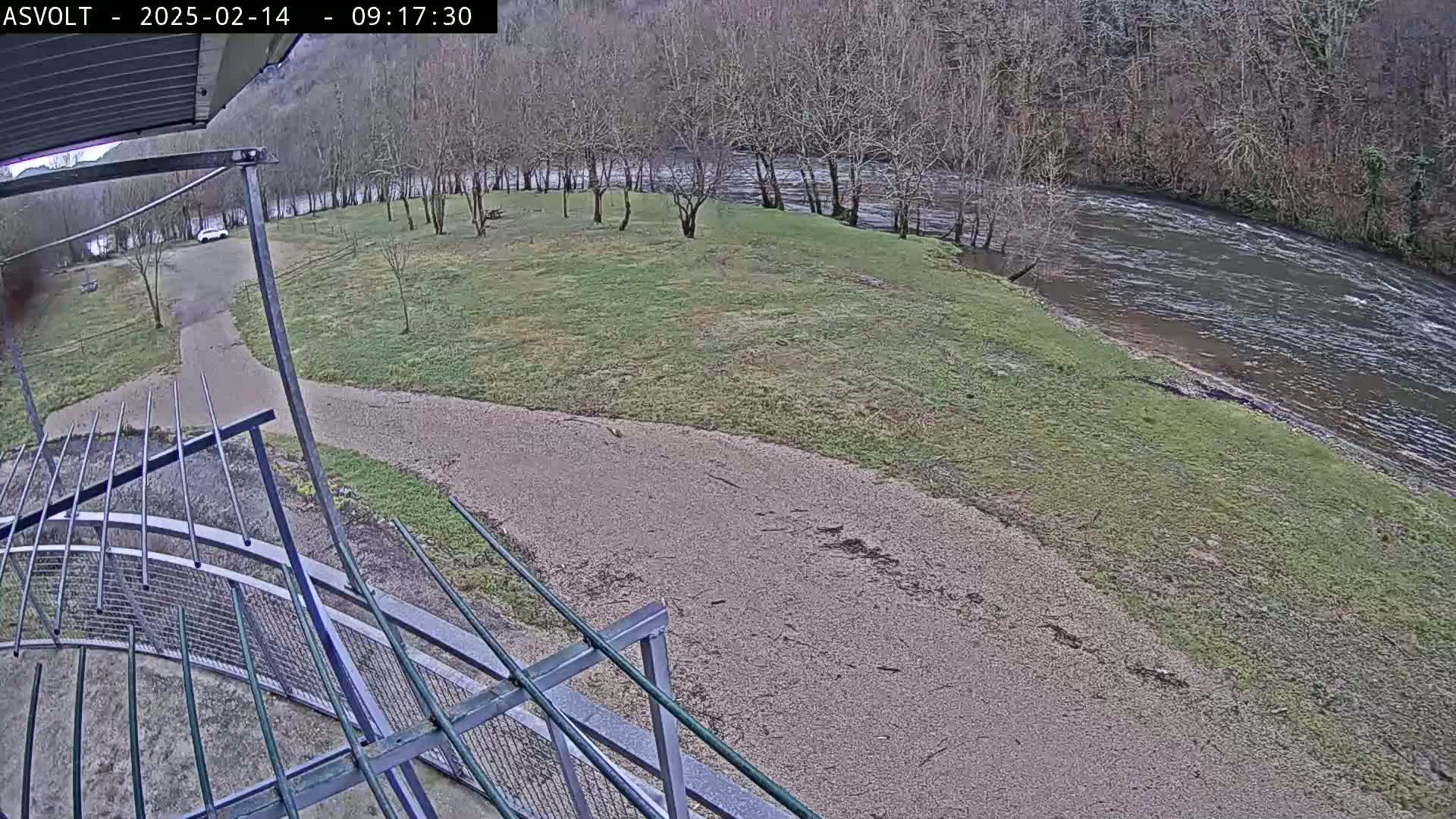 A gravel path leads to a grassy area beside a flowing river, bordered by bare winter trees under an overcast sky.