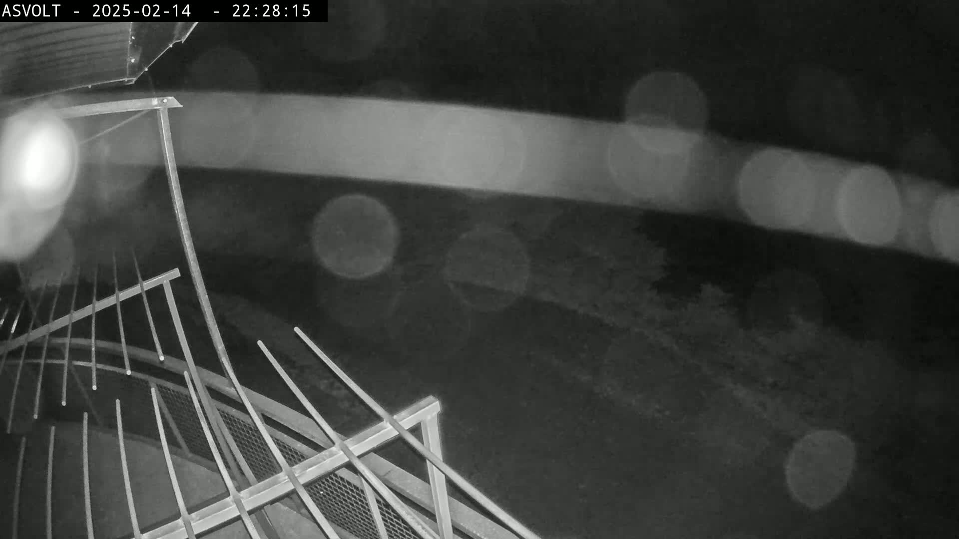 A nighttime, rainy scene shows a metal railing and a blurred, light-streaked background.