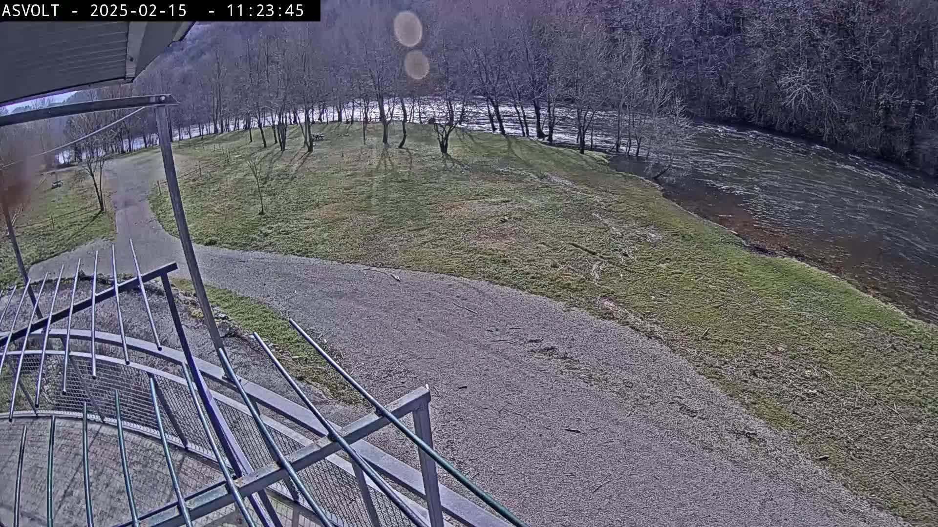 A view from an elevated vantage point shows a gravel path leading to a grassy area beside a river under a partly sunny sky, with bare trees lining the riverbank.