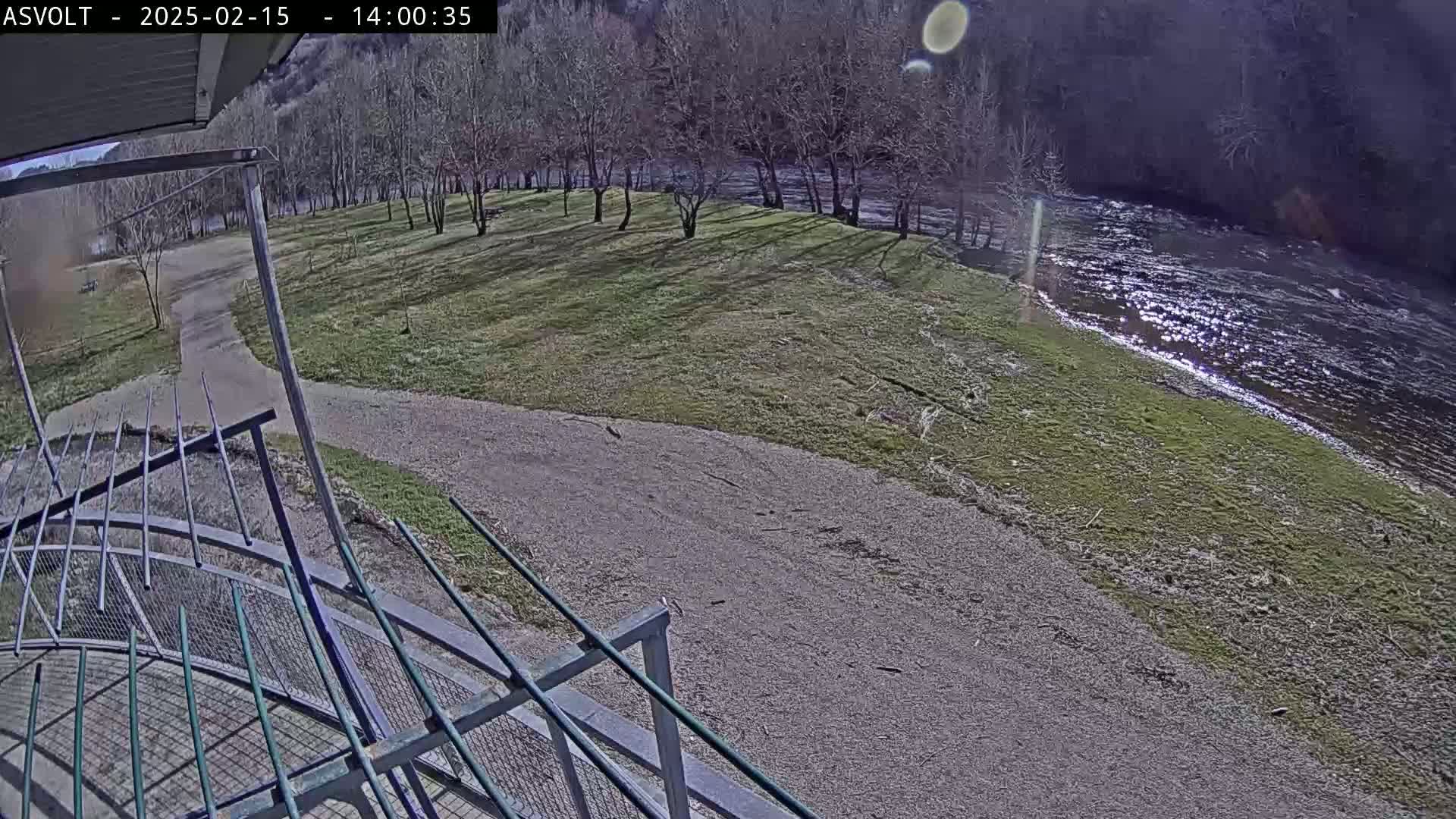 A sunny day shows a gravel path leading to a grassy area beside a flowing river, with bare trees lining the riverbank and a metal railing in the foreground.