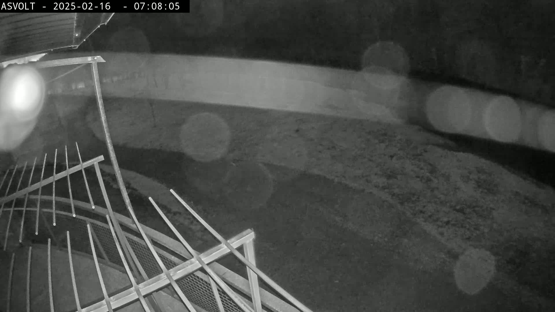A nighttime, low-light view shows a metal railing overlooking a dark, ground-level area with blurry light sources in the background.