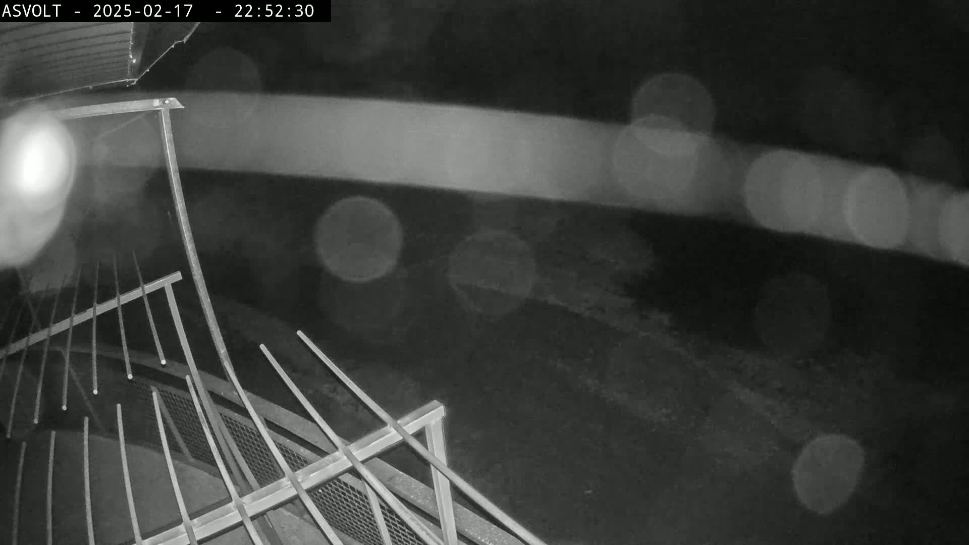 A nighttime, rainy view shows a metal railing and a blurry, indistinct area beyond it.