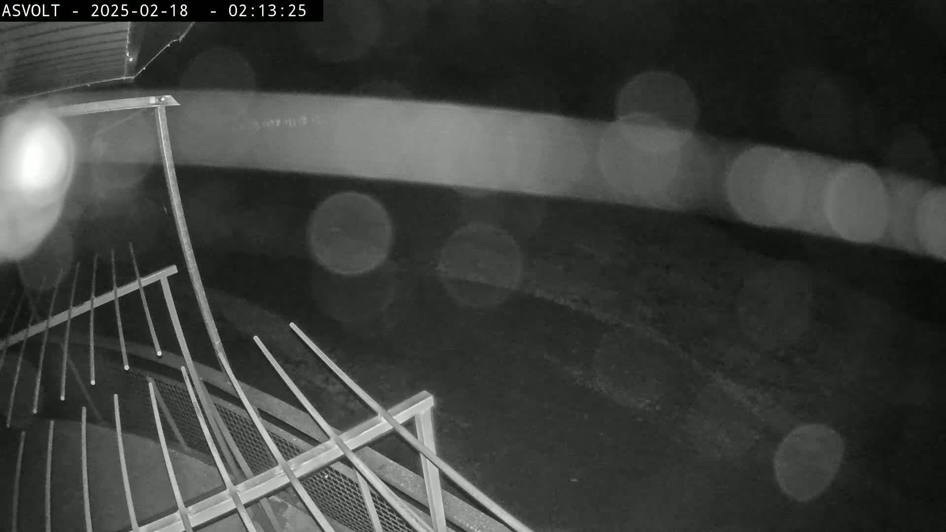 A nighttime, rainy scene shows a metal railing and blurred lights in the background.