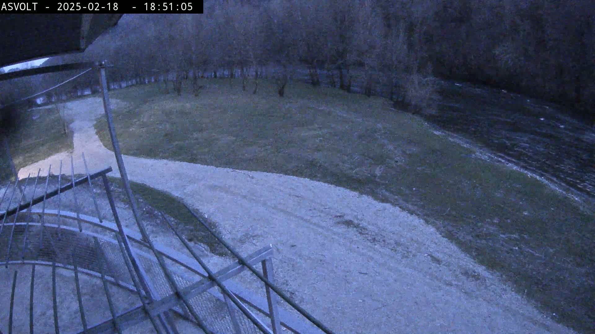 A gravel path curves alongside a riverbank under a twilight sky, bordered by bare trees and a metal railing.