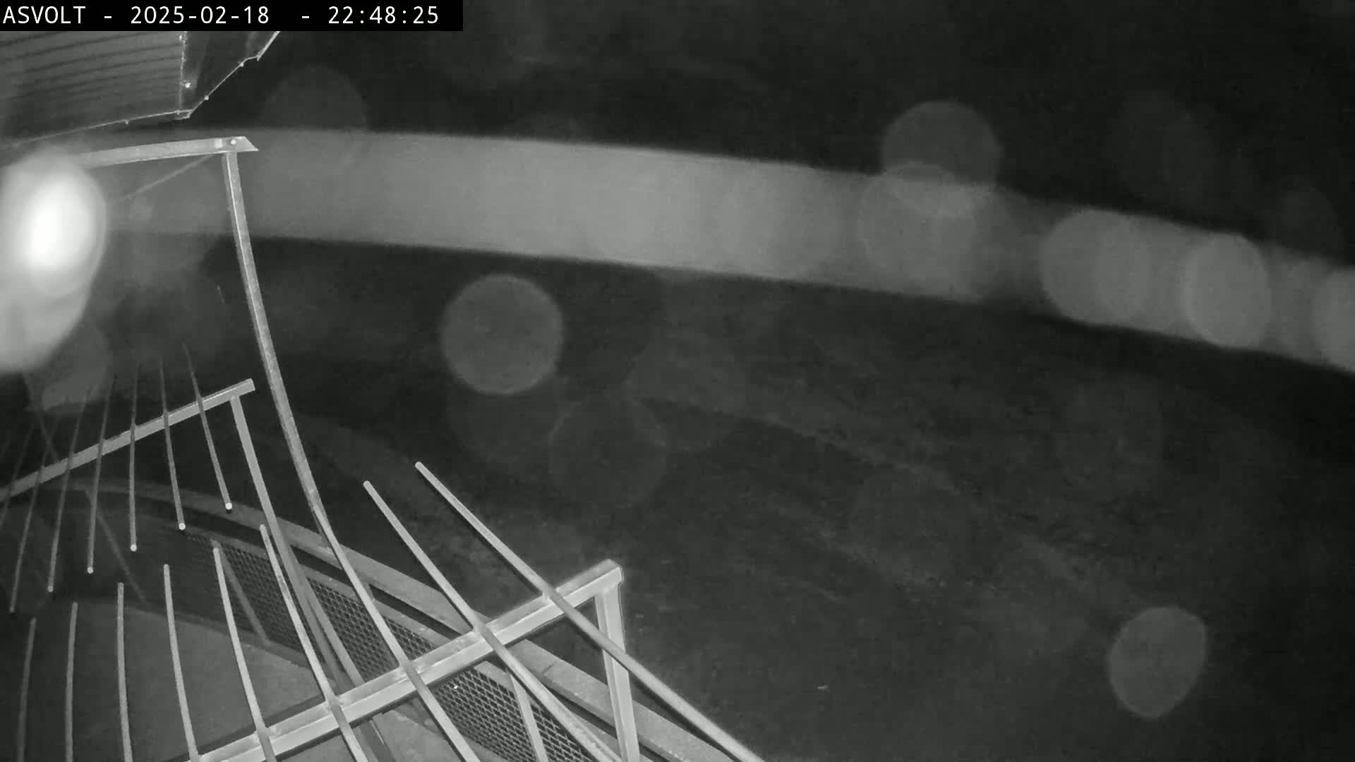 A nighttime, rainy view shows a metal grate or railing in the foreground with blurred lights in the background.