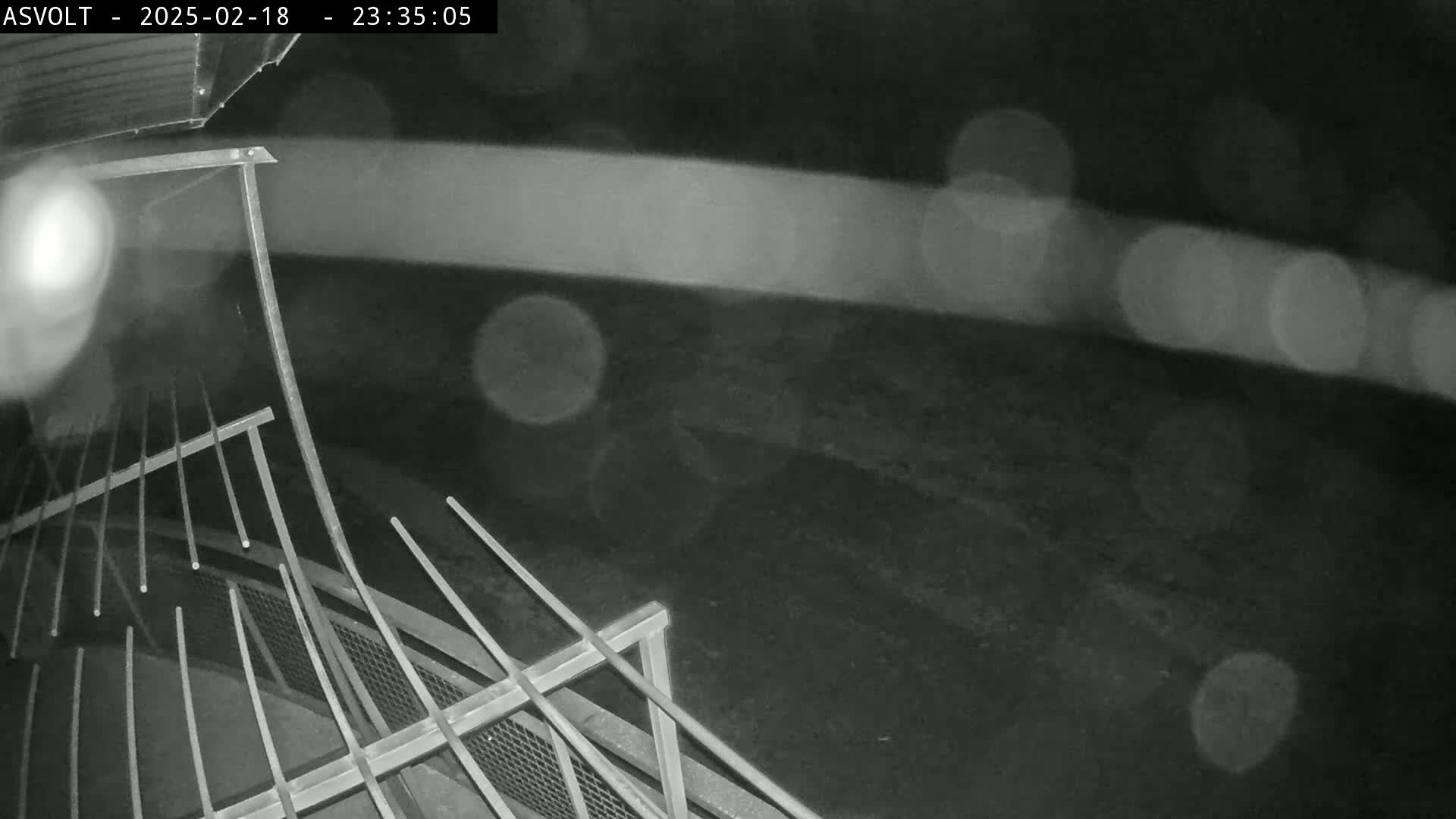 A nighttime, rainy view shows a metal railing and blurry lights in the distance.