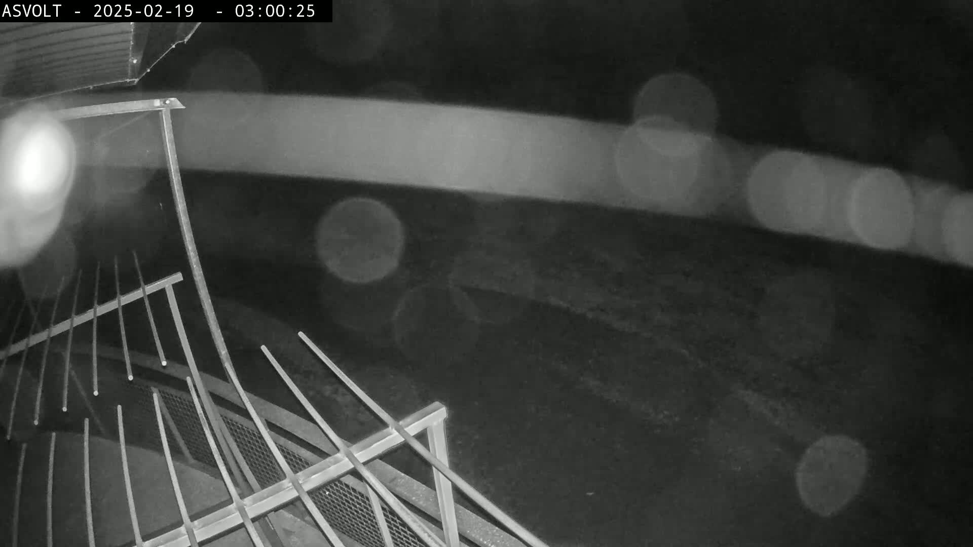 A nighttime, rainy view from a high angle shows a metal railing extending into a dark, blurry background.