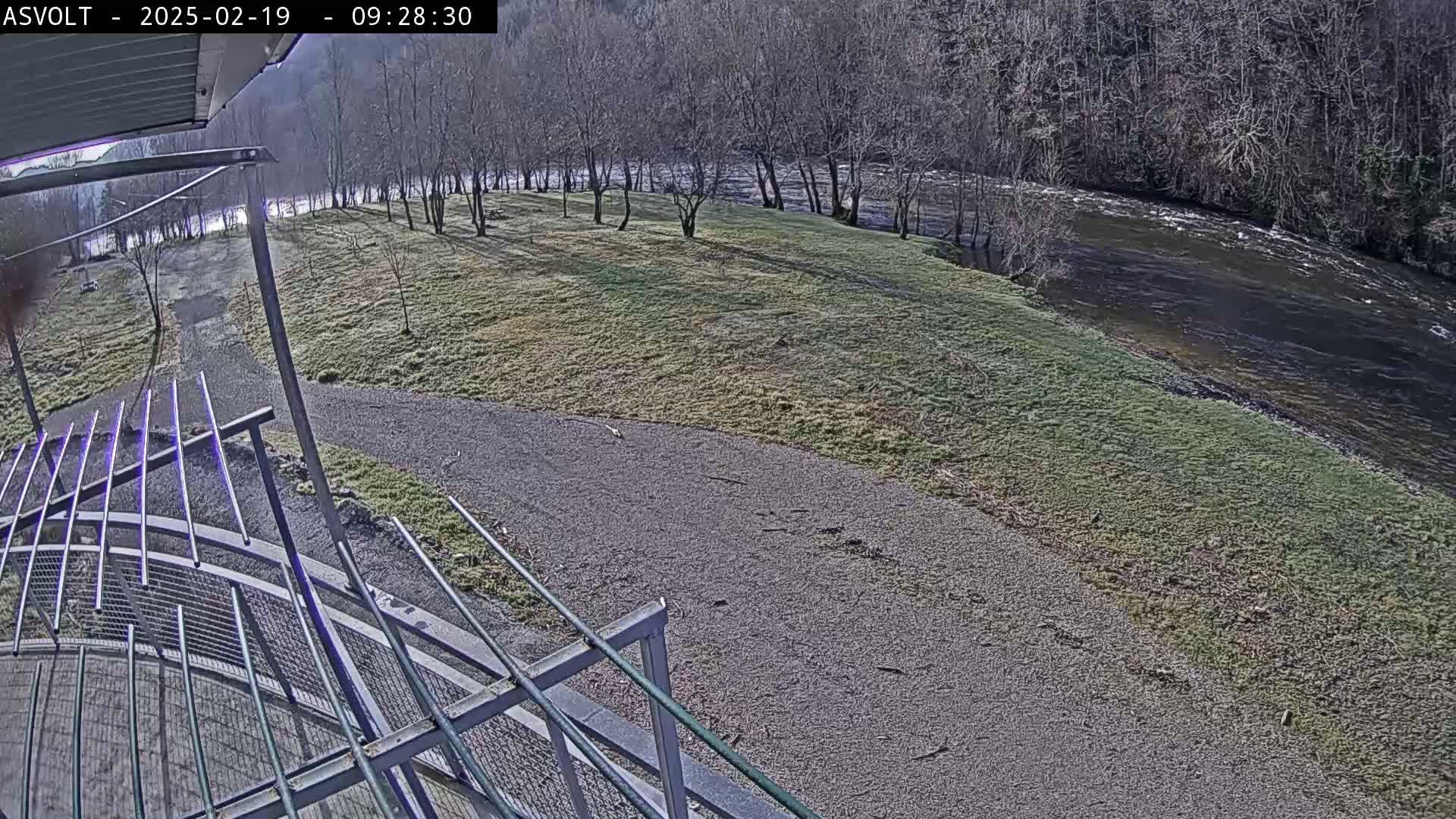 A gravel path leads to a grassy area beside a river under a mostly sunny sky, with leafless trees lining the riverbank.