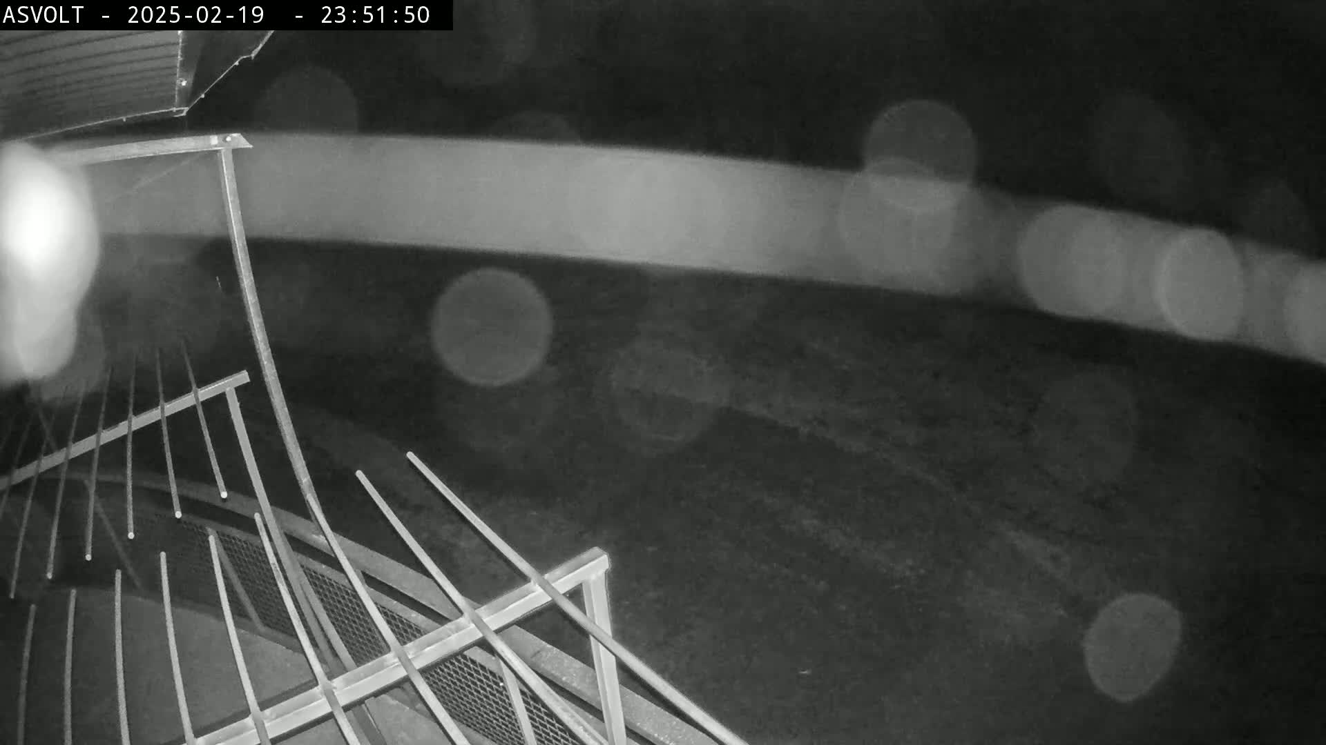 A nighttime, rainy scene shows a metal railing angled toward the viewer, with blurry lights in the background.