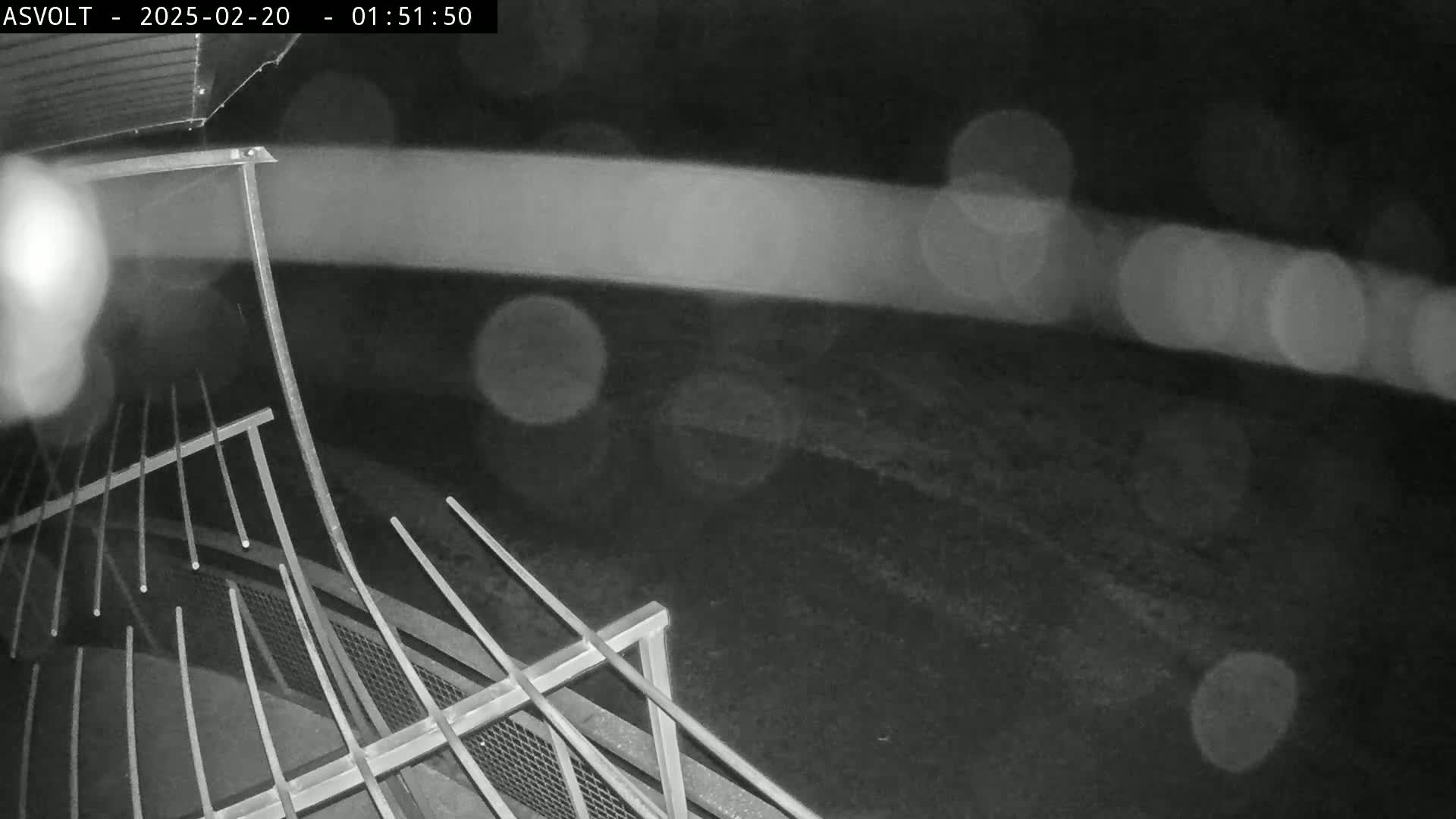 A nighttime, rainy view shows a metal railing and a blurred, dark ground beyond.