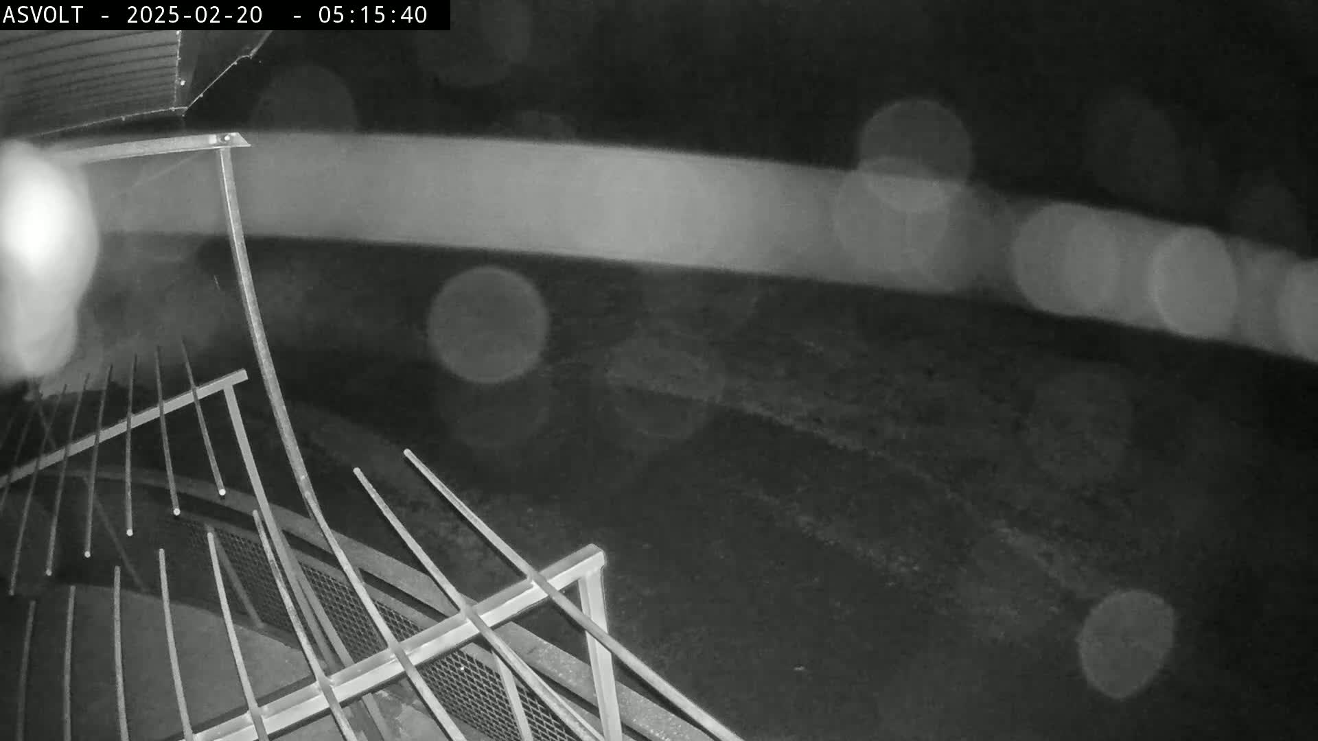 A nighttime, rainy view shows a metal railing and a dark ground in the background.