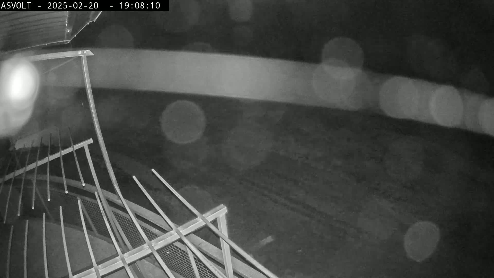 A nighttime, rainy scene shows a metal structure with railings, possibly a portion of a bridge or elevated walkway, overlooking a dark, indistinct ground.