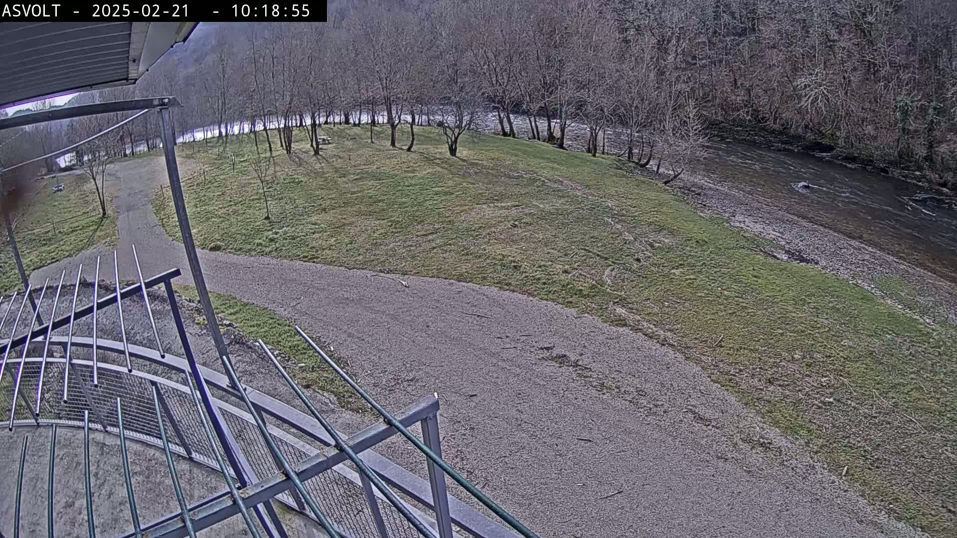 A gravel path winds through a grassy area toward a river bordered by bare trees, under a mostly clear sky.
