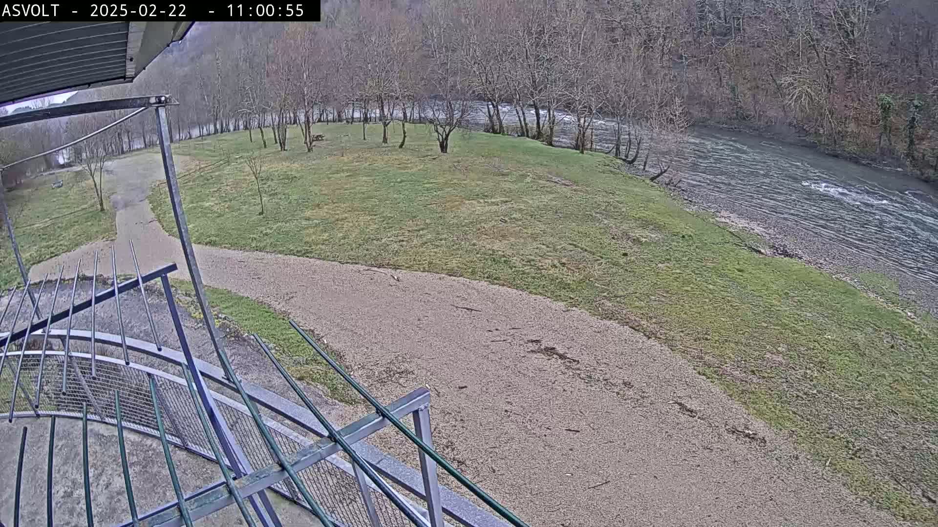 A gravel path curves through a grassy area near a flowing river under an overcast sky.