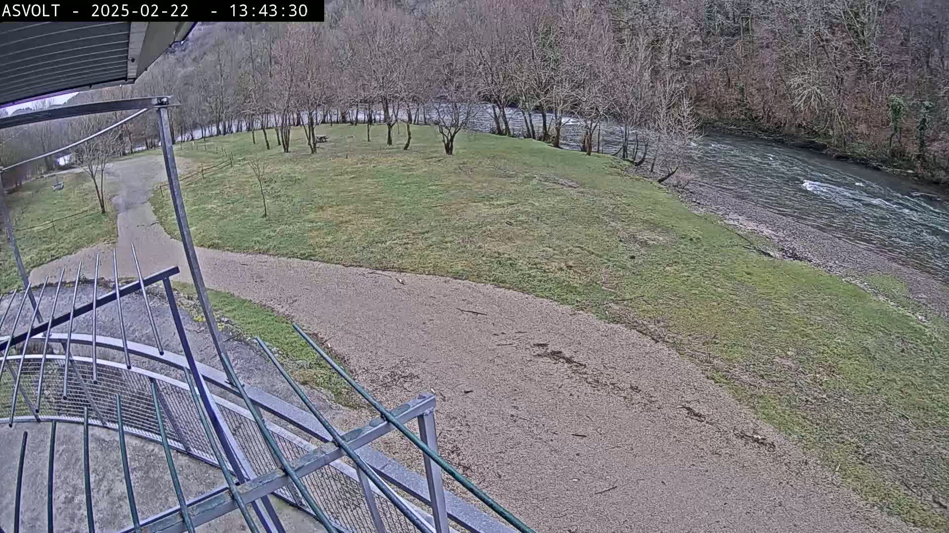A gravel path leads to a grassy area next to a flowing river under an overcast sky.