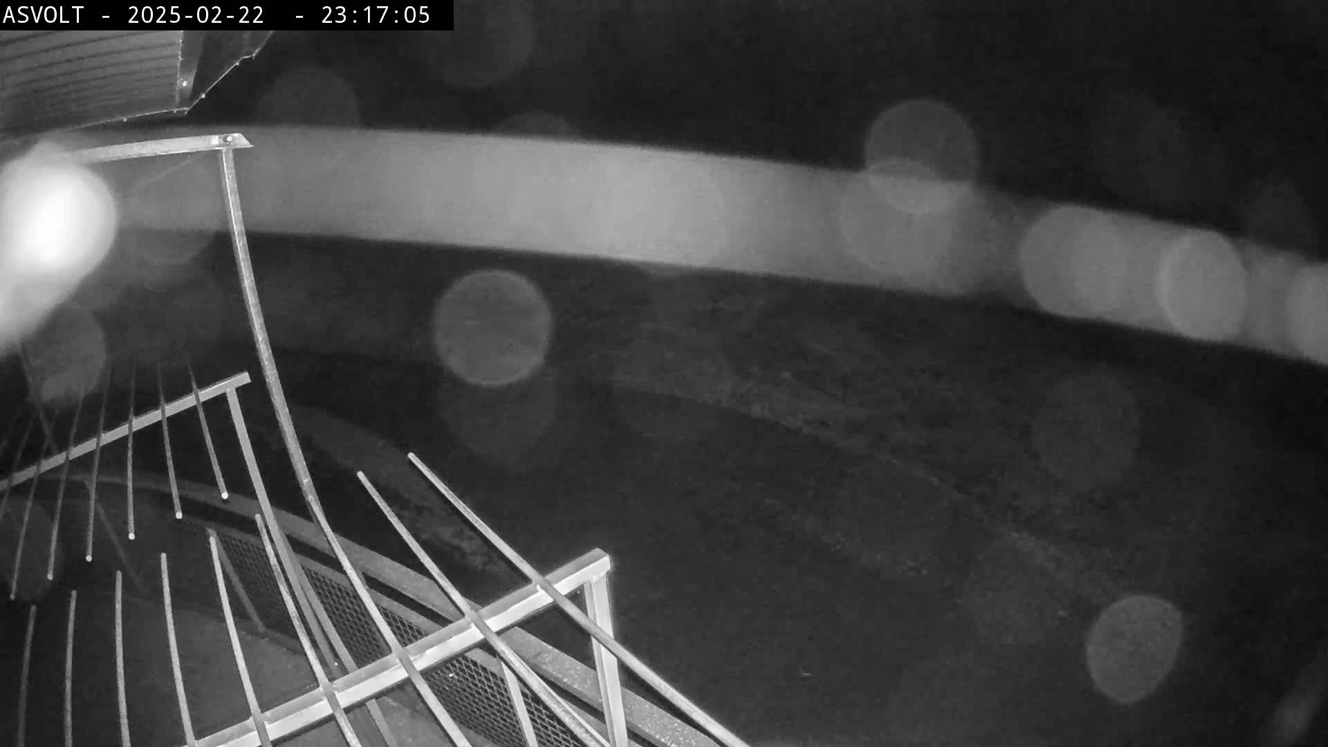 A nighttime, rainy view shows a metal railing and a blurry, indistinct area beyond.