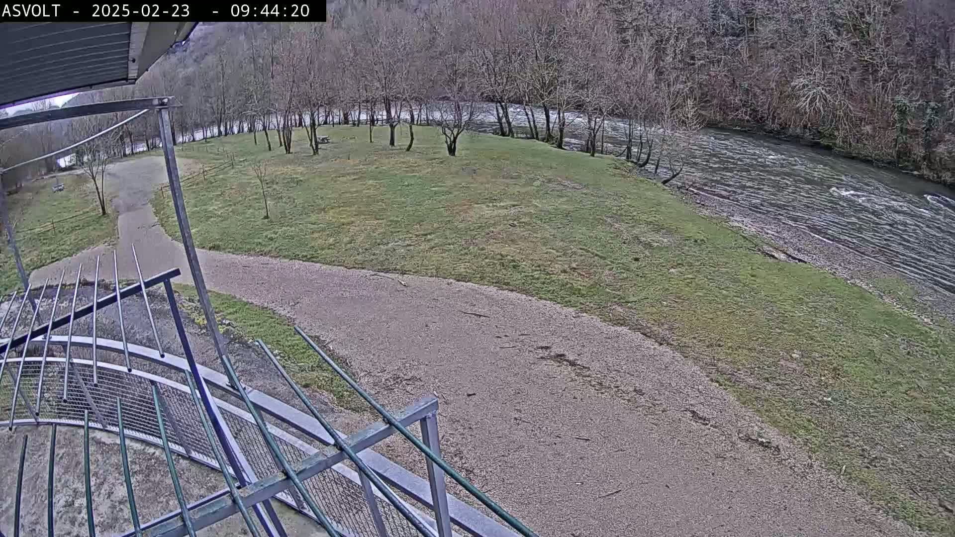 A gravel path curves through a grassy area next to a flowing river, with bare trees lining the riverbank under an overcast sky.