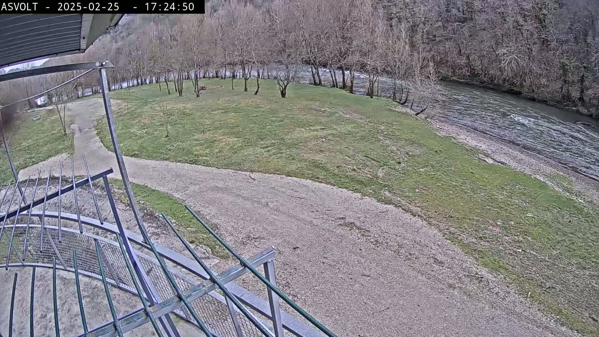 A gravel path leads to a grassy area beside a flowing river, with bare trees lining the riverbank in the background under an overcast sky.