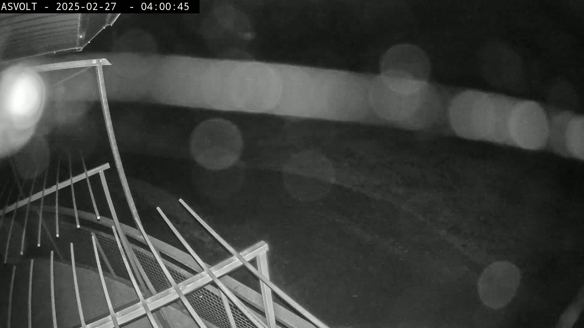 A nighttime, blurry image shows a metal railing in the foreground with blurred lights and a dark ground in the background, possibly rain or moisture present.