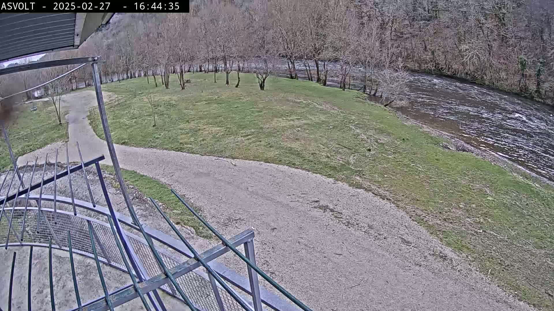 A gravel path curves through a grassy area next to a river, with bare trees lining the riverbank on a cloudy day.